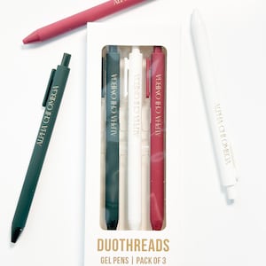 May include: A set of gel pens in a white box, with the text "DUOTHREADS GEL PENS | PACK OF 3". The pens are in dark green, white, and burgundy colors, with the text "ALPHA CHI OMEGA" printed on them. Two additional pens are also visible.