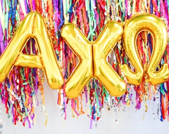 May include: Gold foil letter balloons spelling out "AXO" hanging from a colorful streamer backdrop.