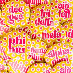 May include: Close-up of several yellow and pink buttons with white stars and sorority names written in pink letters. The names include "Phi Mu", "Tri Delta", "Alpha Gam", "Dee Gee", "Sigma Delta", "Theta", and "Alpha Phi".