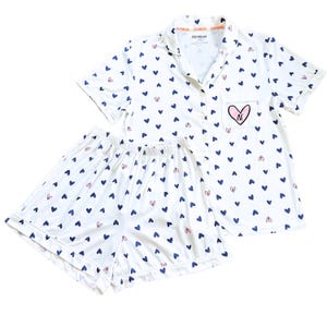 May include: White pajama set with a pattern of blue and pink hearts. The short-sleeved top has a breast pocket with a pink heart design. The shorts have an elastic waistband. The text "DUCTTHREADS" is printed on the collar.