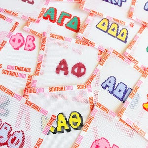 May include: Close-up of several white canvas squares with colorful Greek letters embroidered on them. The letters are in various colors, including red, green, pink, and blue. The squares are surrounded by ribbons with the text "DUOTHREADS".