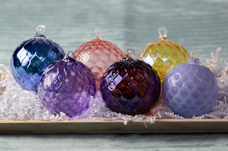May include: Six glass ornaments in various colors including purple, blue, yellow, and pink. The ornaments have a textured, diamond-shaped pattern. The ornaments are displayed in a wooden tray with white shredded paper.