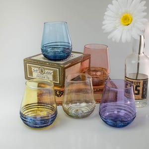 May include: A collection of five stemless drinking glasses in blue, yellow, pink, clear, and purple, each featuring horizontal etched lines near the base. The glasses are arranged around a rectangular box labeled Gold Label Jaguar 70 and a glass bottle.