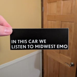 May include: A black rectangular sticker with white text that reads "IN THIS CAR WE LISTEN TO MIDWEST EMO". The sticker is held up by a hand in front of a door and wall.