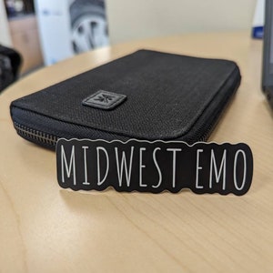 May include: A black zippered wallet with a small logo on the front. The wallet is sitting on a light wood surface with a black sticker that says "MIDWEST EMO" on it.