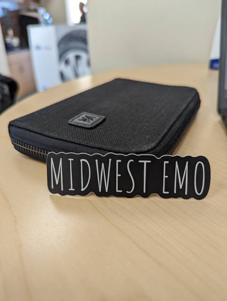 May include: A black zippered wallet with a small logo on the front. The wallet is sitting on a light wood surface with a black sticker that says "MIDWEST EMO" on it.