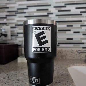 May include: A black Yeti tumbler with a silver lid and base. The tumbler has a white sticker with the text "Rated E for Emo".