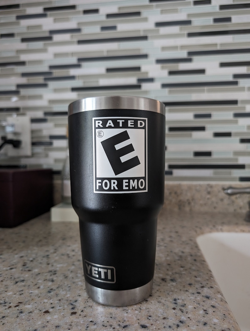 May include: A black Yeti tumbler with a silver lid and base. The tumbler has a white sticker with the text "Rated E for Emo".
