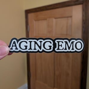 May include: A black and white sticker with the words "AGING EMO" in a bold, sans-serif font. The sticker is held by a person's fingers. The background includes a wooden door and a light-colored wall.