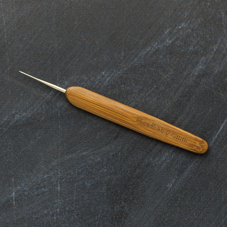 May include: A bamboo dreadlock crochet hook with a silver metal hook tip. The text "DreadLab 0.75mm" is etched into the wooden handle. The hook is designed for creating and maintaining dreadlocks.