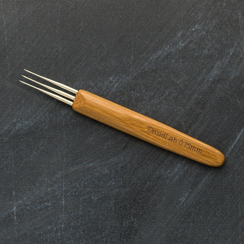 May include: A bamboo dreadlock tool with three metal prongs. The handle is light brown with the text "DreadLab 0.75mm" etched into the wood. The tool is designed for creating and maintaining dreadlocks.
