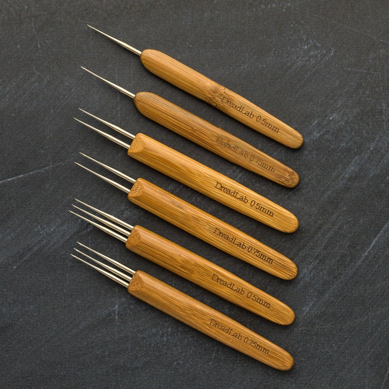 May include: A collection of six bamboo dreadlock tools, each with a silver metal hook. The tools are labeled with "DreadLab" and size markings of 0.5mm and 0.75mm. The tools are arranged diagonally on a dark gray surface.