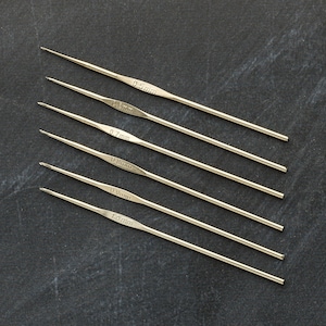 Peut inclure: Un ensemble de six aiguilles à tricoter argentées sur une surface gris foncé. Les aiguilles sont étiquetées avec leurs tailles en millimètres : 0,5 mm, 0,6 mm, 0,7 mm, 0,8 mm, 0,9 mm et 1,0 mm.