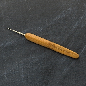 May include: A close-up of a bamboo dreadlock tool with a metal tip. The tool has the text "DreadLab 0.5mm" etched into the bamboo handle. The tool is set against a dark gray background.