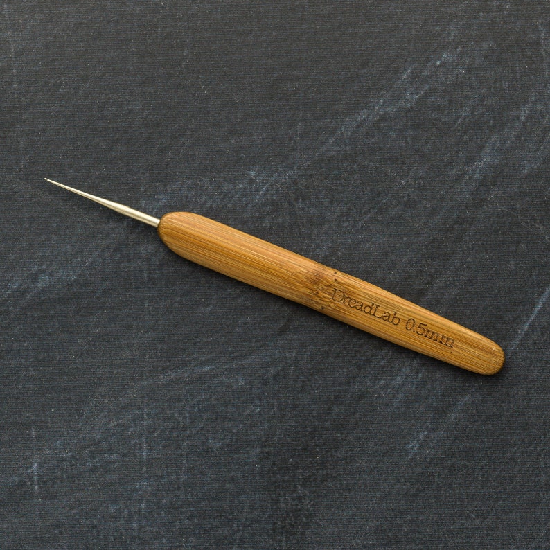 May include: A close-up of a bamboo dreadlock tool with a metal tip. The tool has the text "DreadLab 0.5mm" etched into the bamboo handle. The tool is set against a dark gray background.