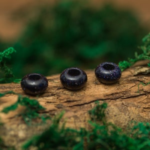 Puede incluir: Tres cuentas de piedra preciosa azul oscuro con textura moteada, dispuestas en fila sobre un trozo de madera cubierto de musgo verde.
