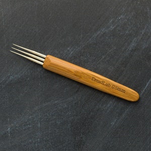 May include: A bamboo dreadlock tool with three metal prongs. The handle is engraved with "DreadLab 0.5mm". The tool is designed for creating and maintaining dreadlocks. The tool is set against a dark, textured background.