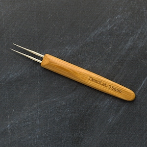 May include: A bamboo dreadlock tool with two silver needles. The tool has the text "DreadLab 0.5mm" engraved on the handle. The tool is designed for creating and maintaining dreadlocks.
