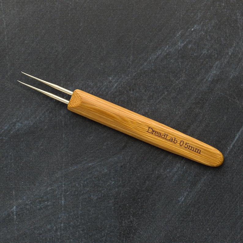 May include: A bamboo dreadlock tool with two silver needles. The tool has the text "DreadLab 0.5mm" engraved on the handle. The tool is designed for creating and maintaining dreadlocks.