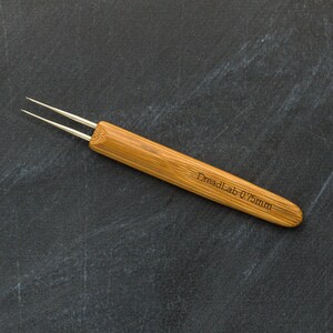 May include: A close-up of a dreadlock tool with a bamboo handle and two silver needles. The text "DreadLab 0.75mm" is etched on the handle. The tool is designed for creating and maintaining dreadlocks.