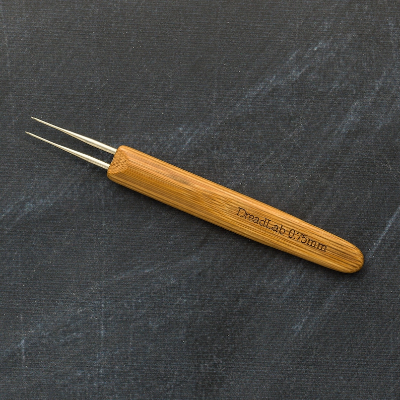May include: A close-up of a dreadlock tool with a bamboo handle and two silver needles. The text "DreadLab 0.75mm" is etched on the handle. The tool is designed for creating and maintaining dreadlocks.