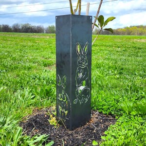 Op de afbeelding: Een zwarte metalen boombeschermer met een uitgesneden ontwerp van twee konijnen. De beschermer is geïnstalleerd rond een jonge boom in een grasveld.