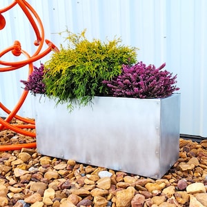 May include: A silver metal planter with three different types of plants. The planter is sitting on a bed of small brown stones. The plants are a light green bush, a dark green bush, and a pink flowering bush.