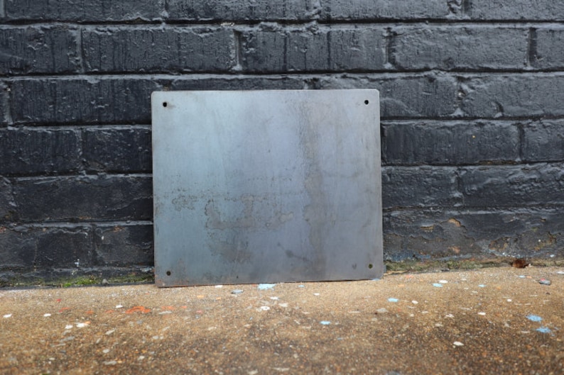 K&ouml;nnte beinhalten: Ein schlichtes, silbernes Metallschild mit abgerundeten Ecken und zwei kleinen L&ouml;chern in den oberen Ecken. Das Schild liegt auf einer Betonfl&auml;che.