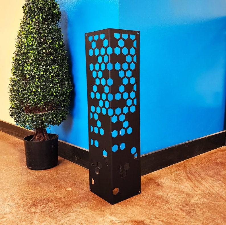 May include: A black metal decorative pillar with a geometric honeycomb pattern and blue backing. The pillar is rectangular and stands upright on a brown floor. A green topiary tree in a black pot is to the left.