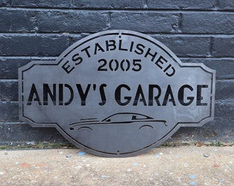 Puede incluir: Un letrero metálico gris oscuro para "Andy's Garage", establecido en 2005. El letrero presenta una silueta de coche y está montado en una pared de ladrillos oscuros. El texto y el diseño del coche están recortados.