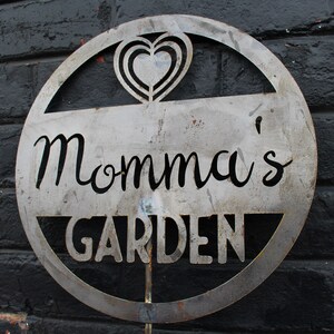 May include: A metal sign with a circle design and the words "Momma's Garden" in a decorative font. The sign has a heart cut out in the top center.