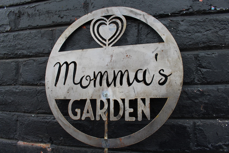 May include: A metal sign with a circle design and the words "Momma's Garden" in a decorative font. The sign has a heart cut out in the top center.