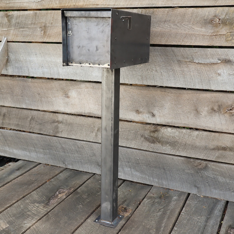 Op de afbeelding: Een zwarte metalen brievenbus met een rechthoekige opening en een klein metalen klepje. De brievenbus is gemonteerd op een hoge, zwarte metalen paal.