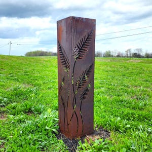 May include: A tall, rectangular, weathered metal garden sculpture. The sculpture features cut-out fern designs. The metal has a rusty brown color, and the sculpture is set in a grassy area under a cloudy sky.