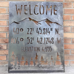 May include: A metal sign with the word "WELCOME" cut out at the top, above a mountain range silhouette. Below are coordinates and the elevation 6,955. A forest silhouette is at the bottom.