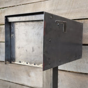 Op de afbeelding: Een zwarte metalen brievenbus met een rechthoekige opening en een metalen klep. De brievenbus is gemonteerd op een metalen paal.