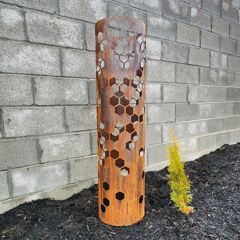 May include: A rusty metal cylinder with a honeycomb pattern cut out. The cylinder is standing on a bed of black mulch.