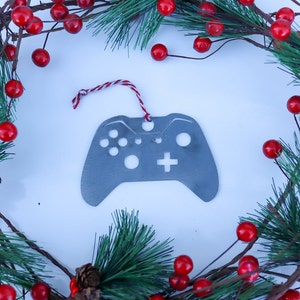 May include: A silver-colored metal ornament shaped like a video game controller, with a red and white string for hanging. The ornament is surrounded by a Christmas wreath of red berries, pine needles, and brown branches.