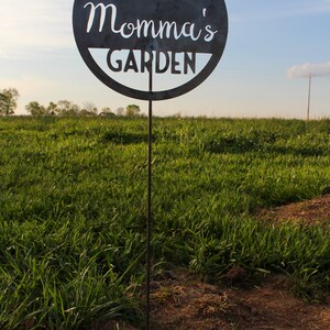 May include: A black metal garden sign with the text "Momma's Garden" and a heart design. The sign is attached to a metal stake and is standing in a field of green grass.