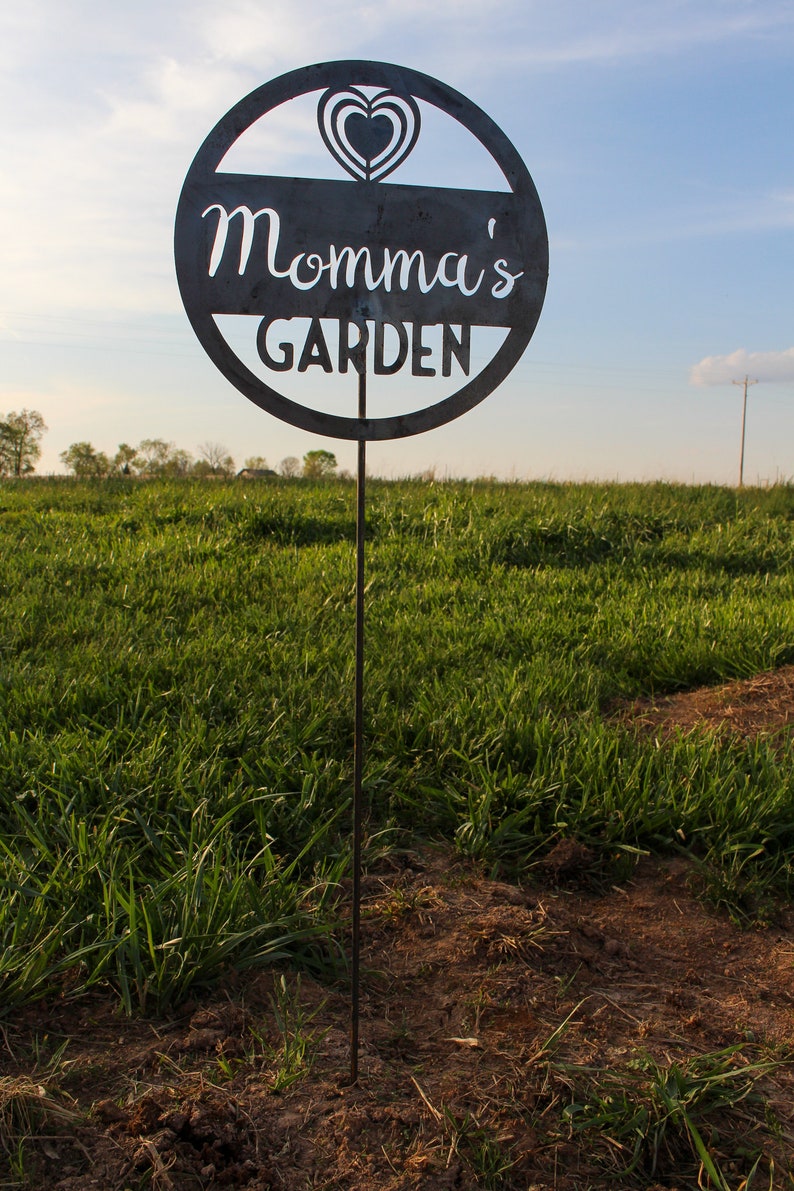 May include: A black metal garden sign with the text "Momma's Garden" and a heart design. The sign is attached to a metal stake and is standing in a field of green grass.