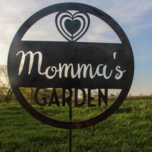 May include: A black metal garden sign with a heart design and the text "Momma's Garden".