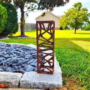 May include: A weathered, rectangular metal garden ornament with a cut-out, abstract pattern. The sculpture is placed on a stone border next to a bed of dark grey stones and green grass. A house and trees are visible in the background.