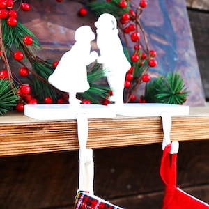 May include: White Christmas stocking holders featuring silhouettes of a boy and girl kissing. The holders are mounted on a wooden shelf, with a red and green Christmas wreath in the background. A red and plaid stocking hangs below.