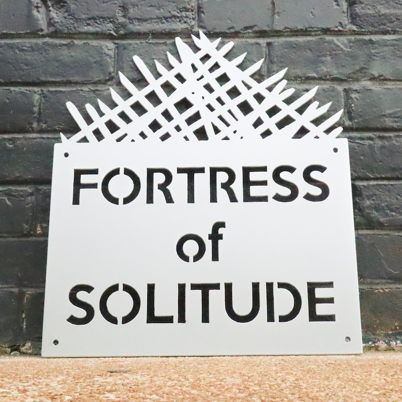 May include: White metal sign with black lettering that reads "FORTRESS of SOLITUDE". The sign has a decorative design at the top that resembles a fence.