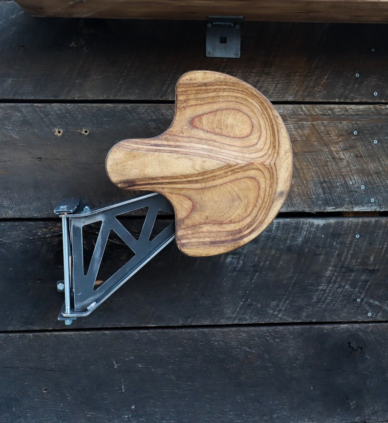 Puede incluir: Un taburete plegable de madera montado en la pared con un soporte met&aacute;lico triangular. El taburete est&aacute; hecho de madera marr&oacute;n claro con un patr&oacute;n de veta natural. El soporte es de metal negro y tiene un dise&ntilde;o geom&eacute;trico.