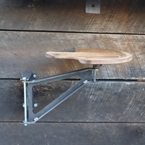 May include: A wooden shelf with a metal bracket attached to a wooden wall. The shelf is curved and has a natural wood finish. The bracket is black and has a triangular shape.