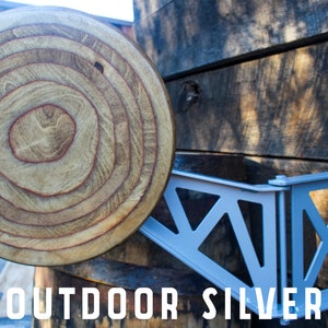May include: A silver metal shelf bracket with a decorative design is mounted to a wooden wall. The bracket is attached to a wooden round disc with concentric rings.