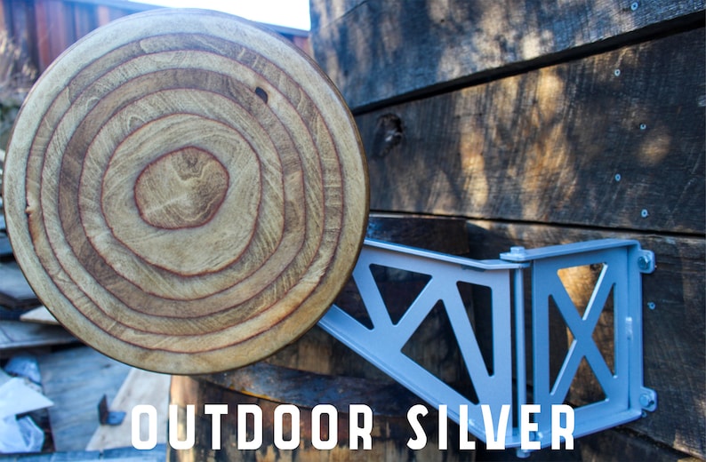 May include: A silver metal shelf bracket with a decorative design is mounted to a wooden wall. The bracket is attached to a wooden round disc with concentric rings.