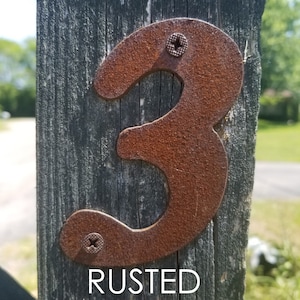 May include: A rusty metal house number three, attached to a wooden fence post. The number is painted in a dark brown color and has a rough, textured surface. The word "RUSTED" is printed below the number.