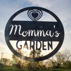 May include: A black metal sign with a circle shape and a heart cut out in the center. The sign says "Momma's Garden" in white lettering.